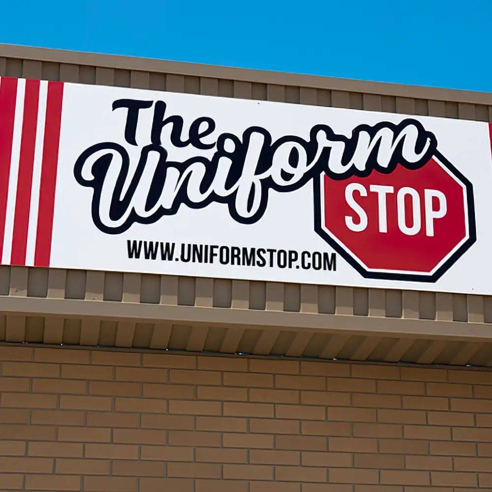 A storefront sign reads The Uniform Stop with STOP inside a red stop sign shape, and the website www.uniformstop.com below, mounted on a tan brick wall. -The Uniform Stop