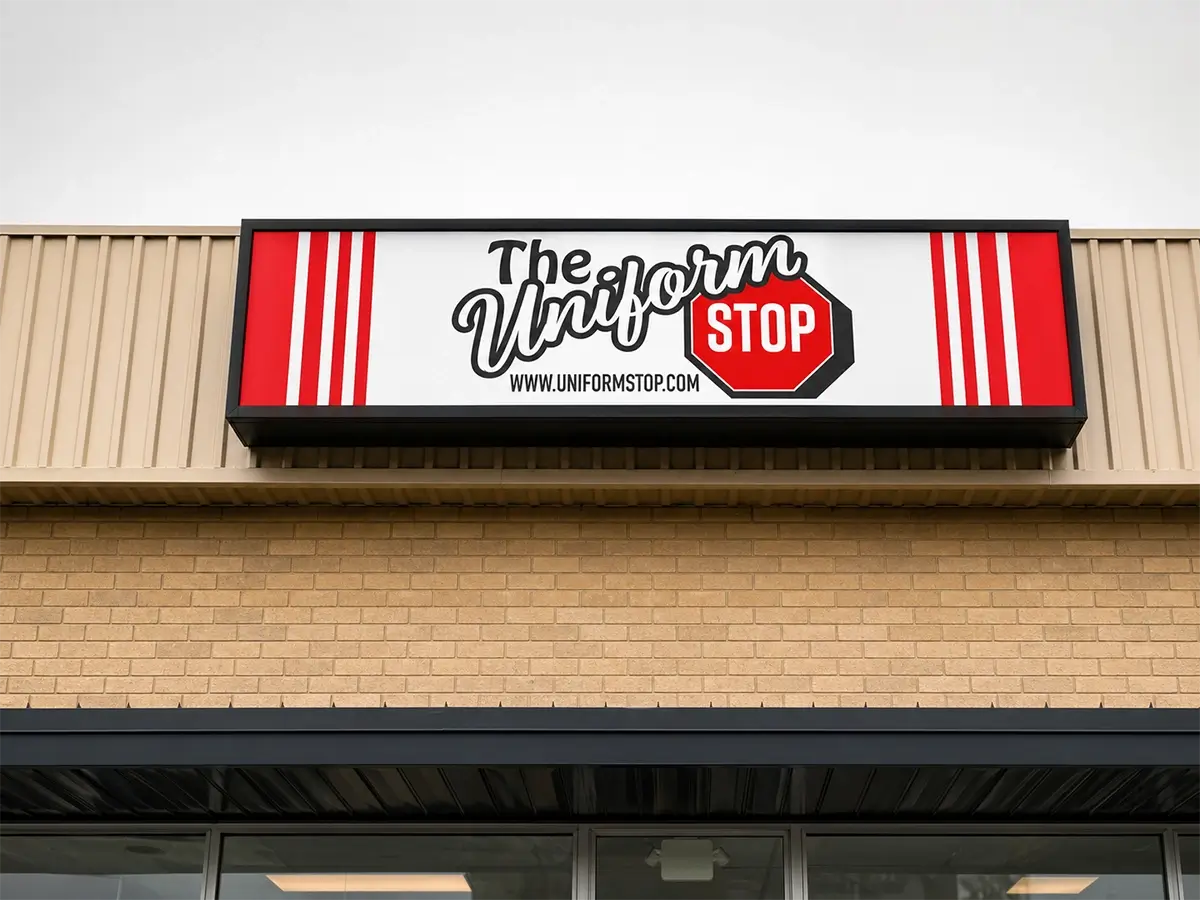 A store sign that reads The Uniform Stop with a red stop sign graphic, bold black and red text, and four vertical red stripes, mounted above a beige brick building. -The Uniform Stop