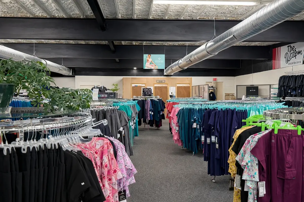 Several racks of medical scrubs in various colors and patterns are displayed in rows inside a well-lit store with exposed ceilings and visible air ducts. A plant and signs can be seen throughout the space. -The Uniform Stop