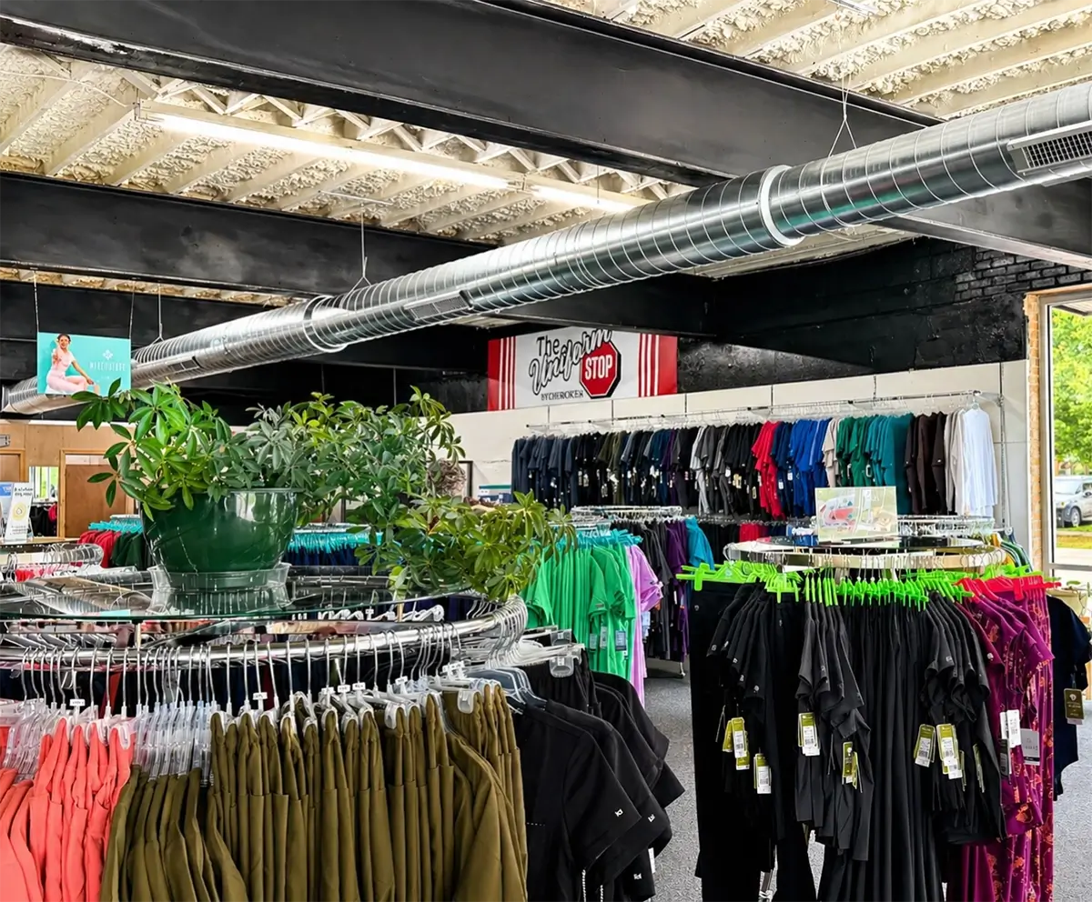 Clothing racks with assorted shirts and pants are displayed inside a thrift store with exposed ceiling pipes and a sign reading The Village Shop Stop above the back wall. A potted plant sits on a circular rack. -The Uniform Stop