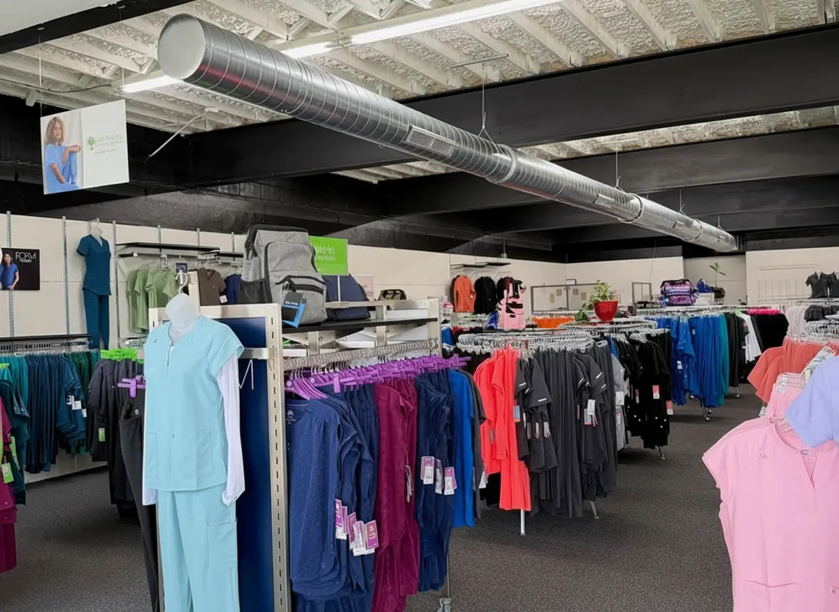 A retail store interior with racks of colorful scrubs and medical uniforms neatly arranged. The ceiling has exposed beams and a silver air duct running across the room. The store is well-lit and organized. -The Uniform Stop