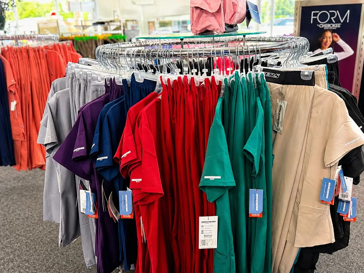 A circular clothing rack displays scrub tops in various colors including purple, navy, red, green, and beige inside a store. Tags are visible on the garments, and other clothes are seen in the background. -The Uniform Stop